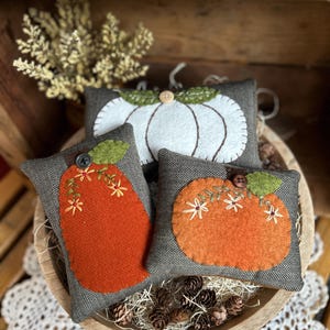 May include: Three rectangular fabric cushions featuring felt pumpkin appliqués. One white pumpkin sits behind two orange pumpkins, all set against a grey herringbone fabric background. Each pumpkin is accented with a green felt leaf, a small button, and embroidered floral details.