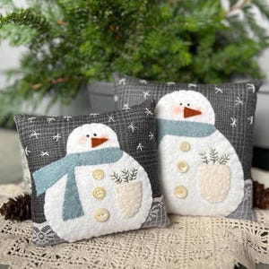 Può includere: Due cuscini decorativi a forma di pupazzo di neve. Ogni cuscino presenta un pupazzo di neve bianco con naso a carota, occhi a bottone e una sciarpa blu. I pupazzi di neve hanno una tasca con rametti verdi e dettagli di bottoni. I cuscini hanno uno sfondo grigio con accenti di fiocchi di neve bianchi.