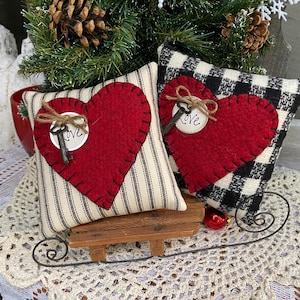 Puede incluir: Dos almohadas de corazón de fieltro rojo con costuras negras y una llave con una etiqueta de "Love" en cada una. Las almohadas están sobre un pequeño trineo de madera y un encaje blanco de ganchillo.