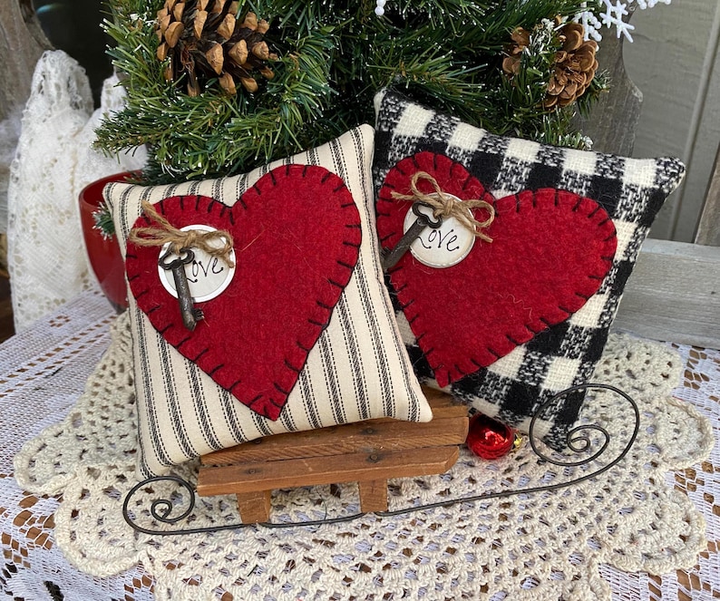 Peut inclure: Deux coussins en forme de c&oelig;ur en feutre rouge avec des coutures noires et une cl&eacute; avec une &eacute;tiquette "Love" sur chacun. Les coussins sont sur un petit tra&icirc;neau en bois et une dentelle blanche au crochet.