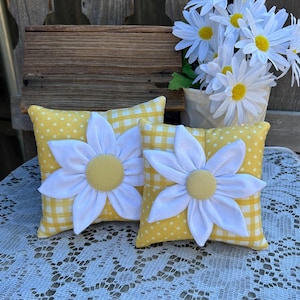 May include: Two decorative throw pillows with a yellow and white daisy design. The pillows have a yellow background with white polka dots and gingham patterns. Each pillow features a large white daisy with a yellow center.