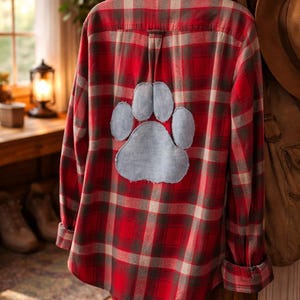 May include: Red and gray plaid flannel shirt with a light blue paw print applique on the back. The shirt is hanging on a wooden wall. A brown hat and jacket are hanging nearby.