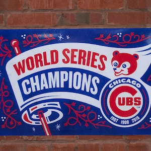 May include: A blue sign with the words "WORLD SERIES CHAMPIONS" in white and red, featuring the Chicago Cubs logo and a cartoon bear. The sign commemorates the team's victories in 1907, 1908, and 2016.