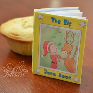 Op de afbeelding: Een klein, geel "The Elf Joke Book" met een cartoonillustratie van een elf en een rendier. Het boek heeft een lichtblauwe rand en sneeuwvlokaccenten. Een gebakken gebakje staat op de achtergrond.