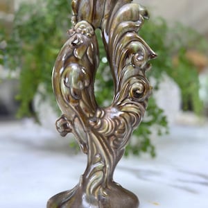 Puede incluir: Figura de gallo de cerámica en tonos marrón y verde. La pieza decorativa presenta diseños detallados de plumas y flores. El gallo se apoya sobre una base redonda y tiene una forma curva de círculo abierto.