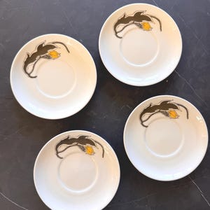 May include: Four circular white ceramic saucers, each featuring a stylized dark olive green leaf design with a small yellow flower accent. The saucers have a central recessed area for a cup and a smooth, glossy finish on the surface.