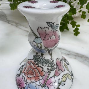 Peut inclure: Bougeoir en céramique blanche avec un motif floral. Le bougeoir présente des fleurs roses, bleues et pêche avec des feuilles vertes. Le dessus du bougeoir a de petits accents de fleurs violettes.