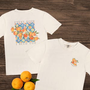 May include: Two white t-shirts with orange fruit graphics. One shirt has a large design with text "EVERY HOUR I NEED YOU". The other shirt has a small orange fruit graphic. Three oranges and a green leaf are on a dark wood surface.