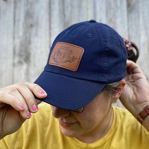 May include: A navy blue baseball cap with a brown leather patch that says "Rooted Truth" and features a floral design. The cap is being worn by a person with a yellow shirt with a sunflower design.