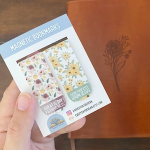 May include: Two magnetic bookmarks with floral designs. One bookmark has a pink and white floral pattern with the text "Great is thy faithfulness". The other bookmark has a white and yellow floral pattern with the text "Turn your eyes upon Jesus".