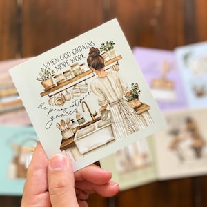 May include: Square art print featuring a kitchen scene with a woman at the sink, surrounded by plants and kitchenware. The print includes the text "When God ordains more work, He pours out more grace." Other prints are visible in the background.