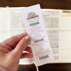 May include: A white bookmark with a colorful Bible study color key. The bookmark features a hand-drawn illustration of an open book with flowers. The key includes categories for truths, commands, promises, and other things to note, such as geography, people, and requests made to God.