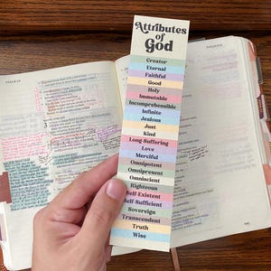 May include: A bookmark with the text "Attributes of God" and a list of attributes in various pastel colors. The bookmark is placed in an open Bible with handwritten notes and tabs.