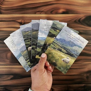 May include: A hand holds a fan of rectangular cards with landscape paintings of green hills, sheep, and mountains. Each card has a different Bible verse printed on it, including "The Lord is my shepherd, I shall not want, Psalm 23:1."
