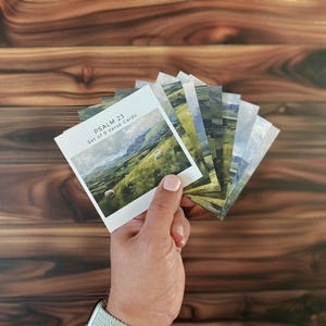 May include: A set of nine verse cards from Psalm 23, featuring landscape images. The cards are held in a hand against a wooden background. The first card displays the text "Psalm 23 Set of 9 Verse Cards."