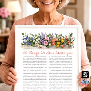 May include: A framed print with a floral arrangement and the text "80 Things We Love About You." The flowers include pink roses, daisies, and other colorful blooms. The print is held by a smiling person.