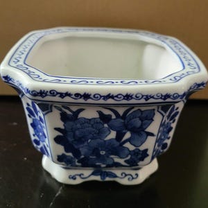 May include: A white ceramic planter with an octagonal shape, featuring blue floral patterns and intricate scrollwork borders. The exterior displays detailed blue painted flowers on each side, while the rim is decorated with a continuous blue line pattern.