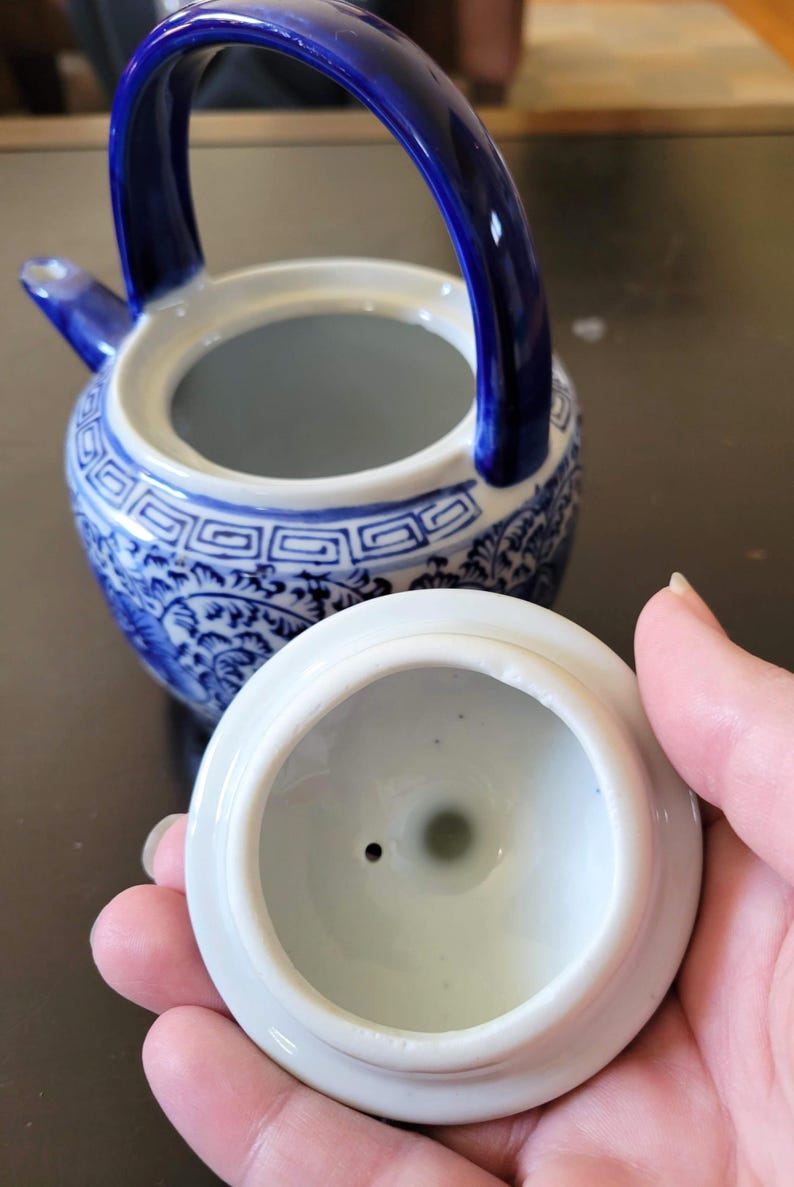 May include: A blue and white porcelain teapot with a lid. The teapot has a floral design and a blue handle. The lid has a small hole in the center.