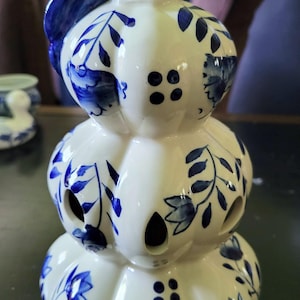 May include: A white ceramic candle holder with a stacked pumpkin design. It features blue floral patterns and cut-out holes for light. The handle and accents are painted in a deep blue color.