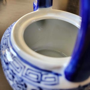 May include: Blue and white porcelain teapot with a blue handle. The teapot has a traditional Chinese design with a geometric pattern and floral motifs. The inside of the teapot is white.
