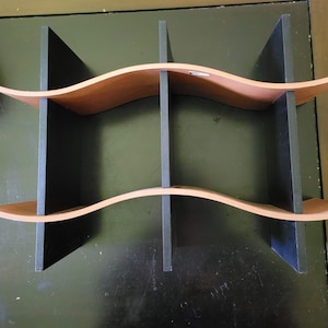 May include: A wooden shelf unit with a wavy design. The shelves are a light brown color, and the dividers are black. The unit has a modern, minimalist aesthetic and is suitable for displaying items.