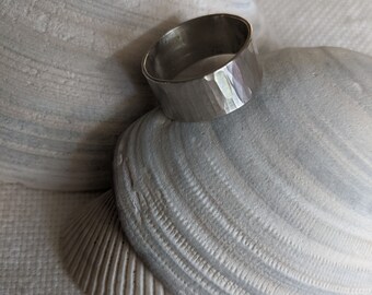 May include: A silver hammered ring with a wide band, resting on a white seashell.