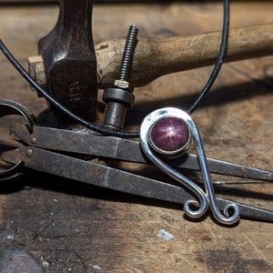 May include: A silver pendant with a pink gemstone set in a bezel setting. The pendant is hanging on a black leather cord. The pendant is lying on a wooden surface with a pair of metal calipers and a hammer.