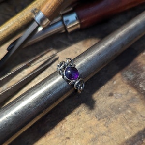 May include: A silver ring with a purple gemstone set in a swirling design.