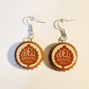 May include: A pair of earrings made from brown bottle caps with the Odell Brewing Co. logo printed on them. The bottle caps are attached to silver earring hooks.