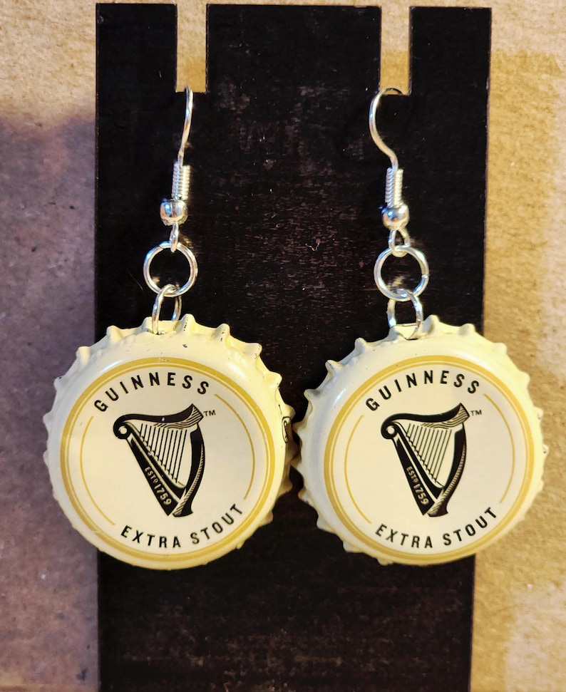 May include: A pair of earrings made from Guinness Extra Stout bottle caps. The earrings feature a white background with the Guinness logo, a black harp, and the words "GUINNESS" and "EXTRA STOUT". The earrings hang from silver-colored hooks.
