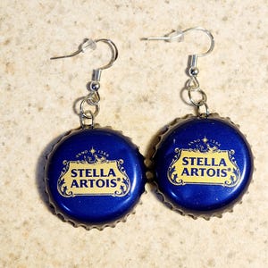 Könnte beinhalten: Ein Paar Ohrringe aus blauen Stella Artois Flaschendeckeln mit goldenen Schriftzügen. Die Ohrringe sind an silbernen Haken befestigt.
