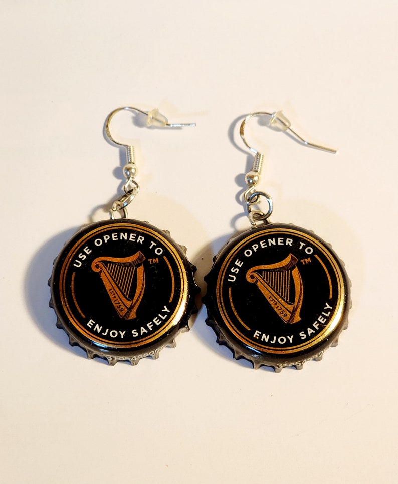 Puede incluir: Un par de pendientes hechos con tapas de botellas de Guinness negras con letras doradas que dicen "USE OPENER TO" y "ENJOY SAFELY" con un logotipo de arpa dorado y los textos "ESTD 1759" y "TM".