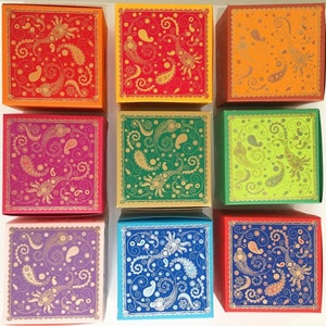 Könnte beinhalten: Neun quadratische Geschenkboxen in verschiedenen Farben, darunter Orange, Rot, Grün und Blau. Jede Box hat ein goldenes Paisley-Design mit komplizierten Wirbeln und einem gewellten Rand. Die Boxen sind in einem Raster angeordnet.