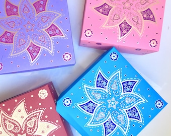 Puede incluir: Cuatro cajas cuadradas con intrincados diseños florales. Las cajas son en tonos de morado, rosa, azul y rojo. Cada caja presenta un patrón floral diferente con acentos dorados.