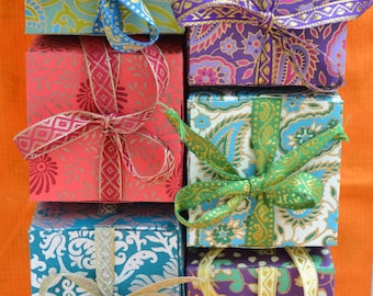 Puede incluir: Seis pequeñas cajas cuadradas de regalo con papel estampado de colores y atadas con lazos de cinta. Las cajas están apiladas en forma de pirámide. Las cajas están decoradas con diferentes colores y patrones, incluyendo azul, verde, morado, rojo y dorado.