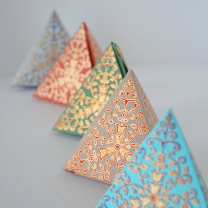 May include: Four triangular boxes with intricate gold and colored patterns. The boxes are arranged in a row, with the largest box in the back and the smallest box in the front. The boxes are made of paper and have a delicate, hand-painted look.