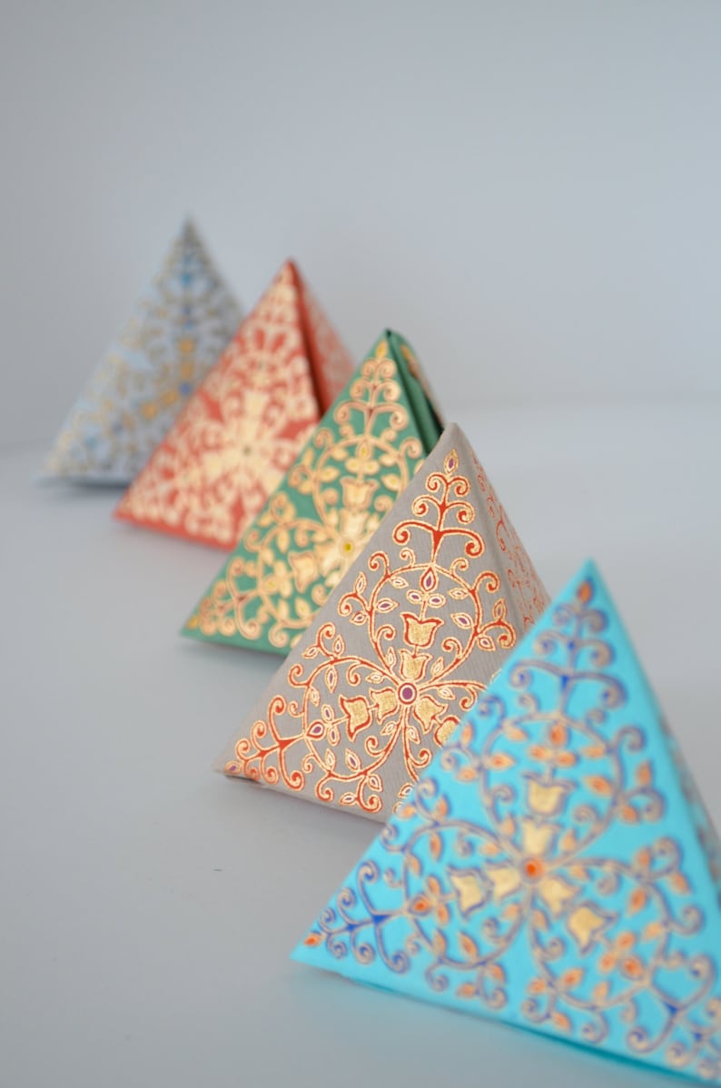 May include: Four triangular boxes with intricate gold and colored patterns. The boxes are arranged in a row, with the largest box in the back and the smallest box in the front. The boxes are made of paper and have a delicate, hand-painted look.