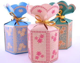 Puede incluir: Tres cajas de regalo hexagonales en azul, rosa y verde azulado, cada una con un diseño floral y una parte superior en forma de pétalo atada con una cinta dorada. Las cajas tienen un patrón decorativo y son adecuadas para pequeños regalos.