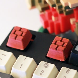 Peut inclure: Un clavier noir avec des touches rouges et des touches blanches. Le clavier a une rangée de touches de fonction étiquetées F2, F3 et F4. Une figurine pixelisée d'un personnage de jeu vidéo se tient derrière le clavier.