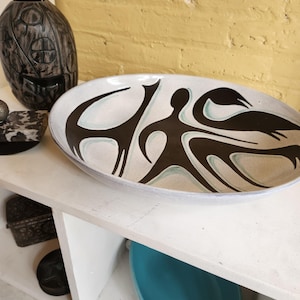 May include: An oval ceramic serving dish with a cream-colored interior and a black and teal abstract design. The dish is displayed on a white shelf, with other decorative items in the background.