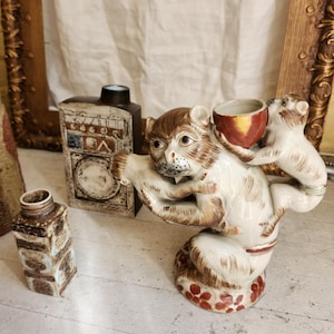 May include: A ceramic figurine of two monkeys, one holding a small red cup. The monkeys are painted in shades of brown, white, and gold. The base of the figurine is decorated with red and white dots.