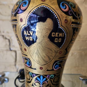 Peut inclure: Un vase décoratif avec un fond noir et des détails dorés. Le vase présente un portrait d'un homme barbu et coiffé d'un chapeau, avec les mots "ALV MO GENI GO" de chaque côté. Le vase présente des motifs floraux colorés.