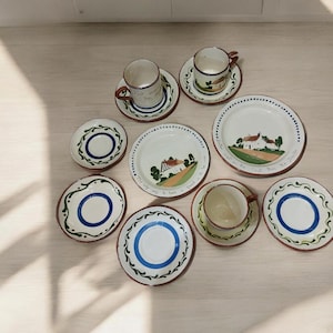 May include: A collection of hand-painted ceramic tableware, including cups, saucers, and plates. The plates feature a white background with a red rim and a blue or green decorative border. Some plates have a painted house scene.