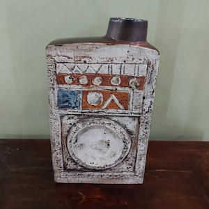 May include: A white ceramic vase with a geometric design in shades of orange, blue, and brown. The vase has a rectangular body and a wide, round opening.