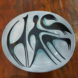 May include: Oval ceramic serving dish with a light blue speckled background. The dish features black abstract figures with teal accents. The figures include a giraffe-like shape, a human-like form, and bird-like shapes.