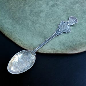 May include: Silver souvenir spoon featuring an engraved design of the Windsor Hotel in Montreal, Quebec, Canada. The handle depicts a man in uniform.