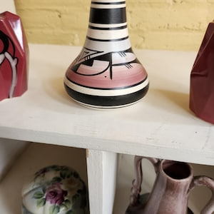 May include: A small, hand-painted ceramic vase with a black, white and pink geometric design. The vase has a rounded base and a narrow neck.
