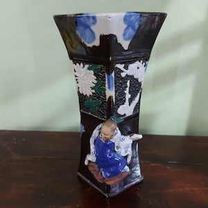 May include: A dark brown ceramic vase with a hexagonal shape and intricate designs. The vase features a white and blue floral pattern, a white and black bird design, and a 3D figure of a person in blue and white clothing.
