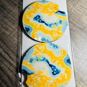 May include: Two car coasters with a vibrant abstract design in yellow, blue, and white. The coasters are round with a curved cutout for easy removal. The packaging reads "Car Coasters" and "Handmade with love."