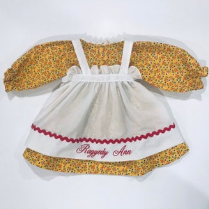 May include: A white dress with red trim and yellow floral print sleeves. The dress has a white apron with the text "Raggedy Ann" embroidered on it.